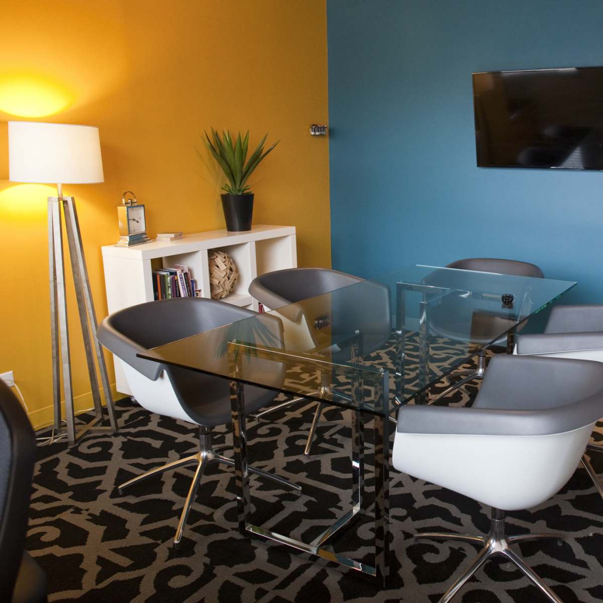 A modern conference room features a glass table surrounded by stylish chairs, with a television mounted on a wall and a lamp beside a plant in one corner.