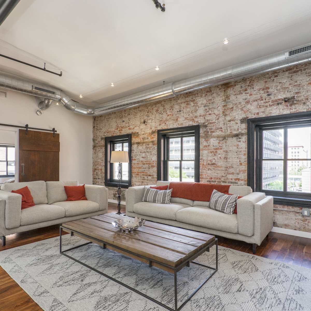 A modern living room with a brick wall, two light-colored sofas, a wooden coffee table, and large windows.