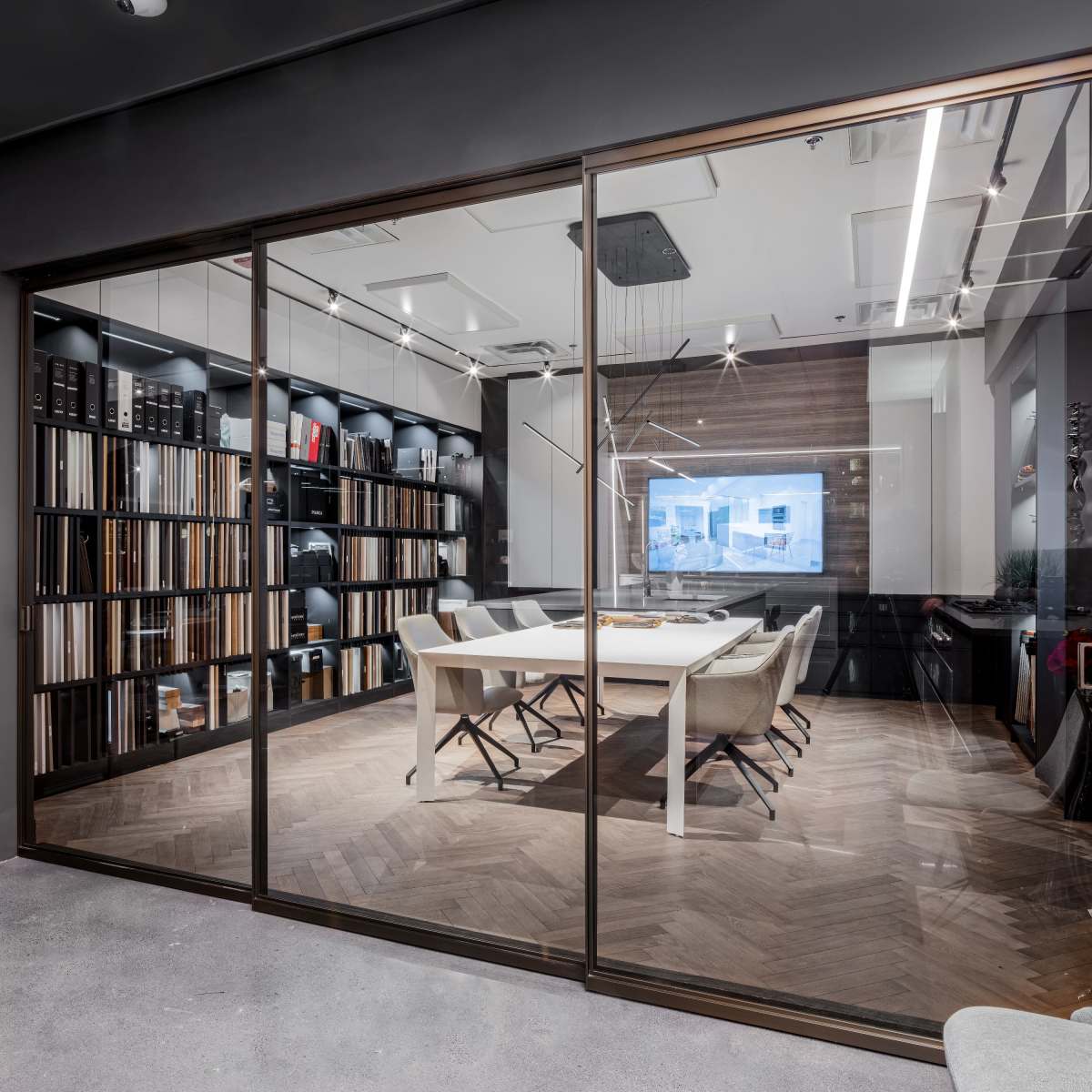 A modern conference room features a glass wall, a large table surrounded by chairs, and shelves filled with design materials.