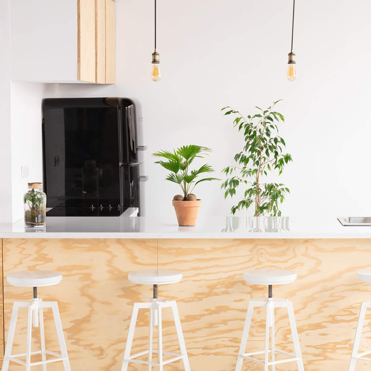A modern kitchen features a light wood countertop with four white stools, a potted plant, and a black refrigerator against a white backdrop.