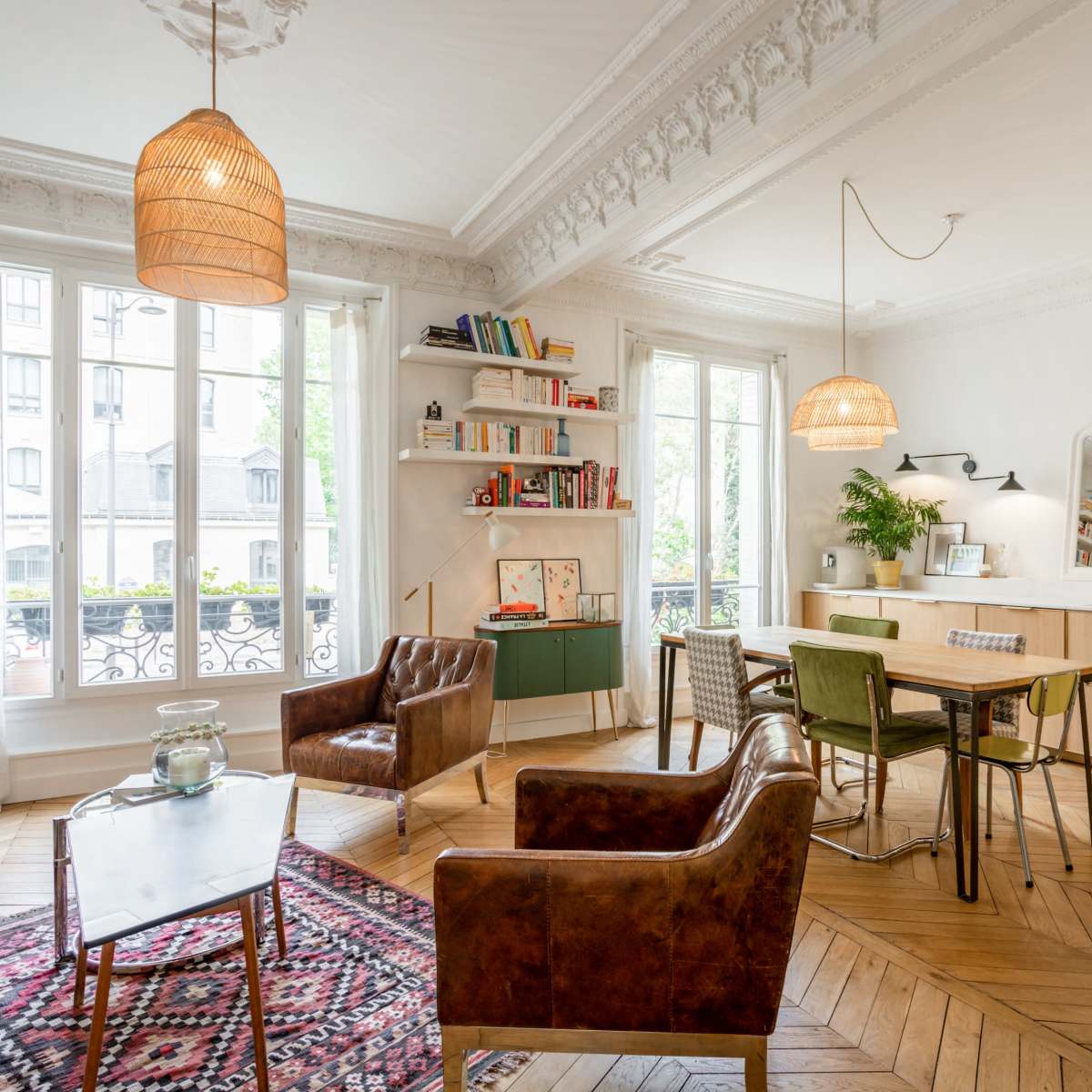 A well-lit living room with a mix of seating, a dining area, bookshelves, and large windows overlooking a street.