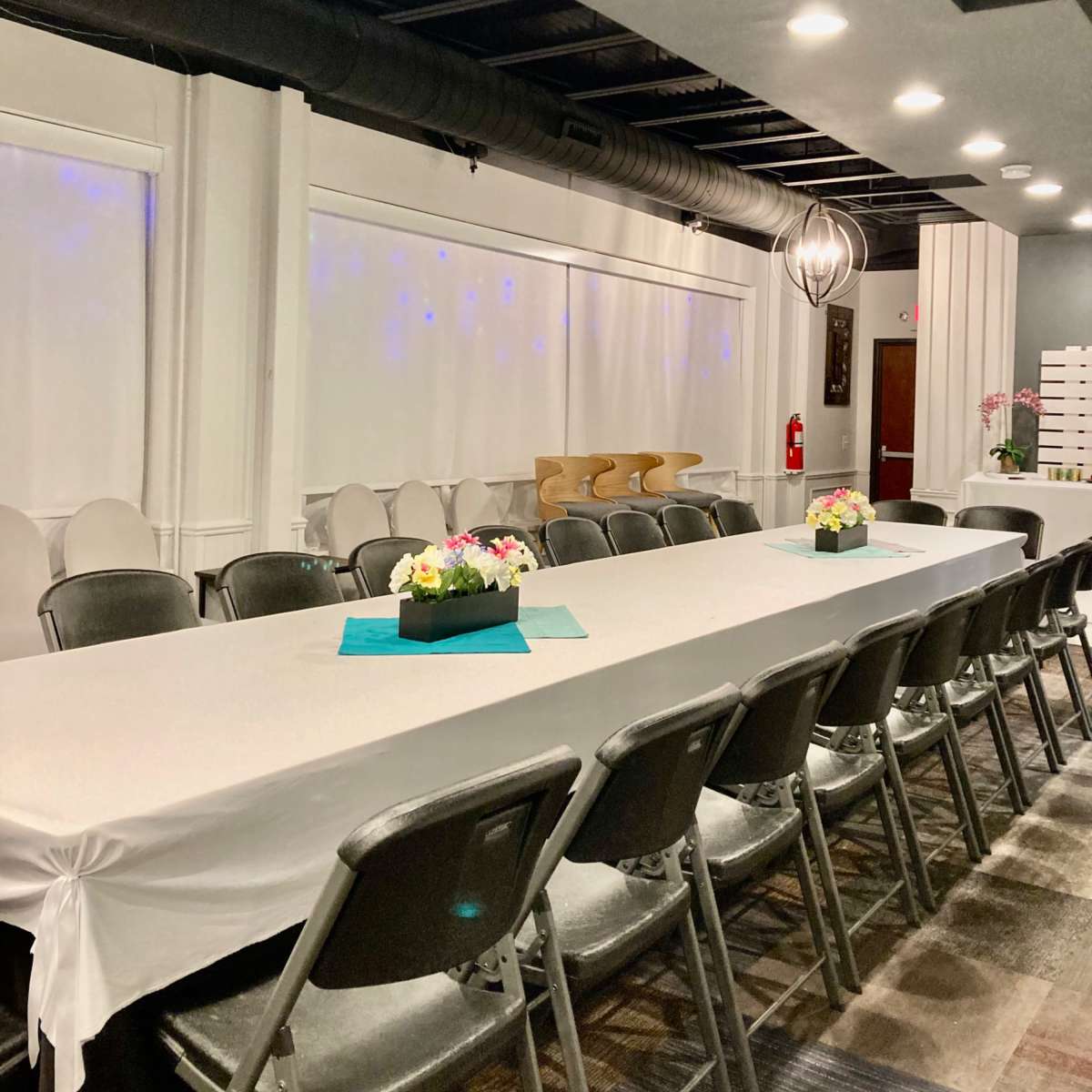 A long, white table with floral centerpieces is set in a spacious room featuring overhead lighting and gray walls.