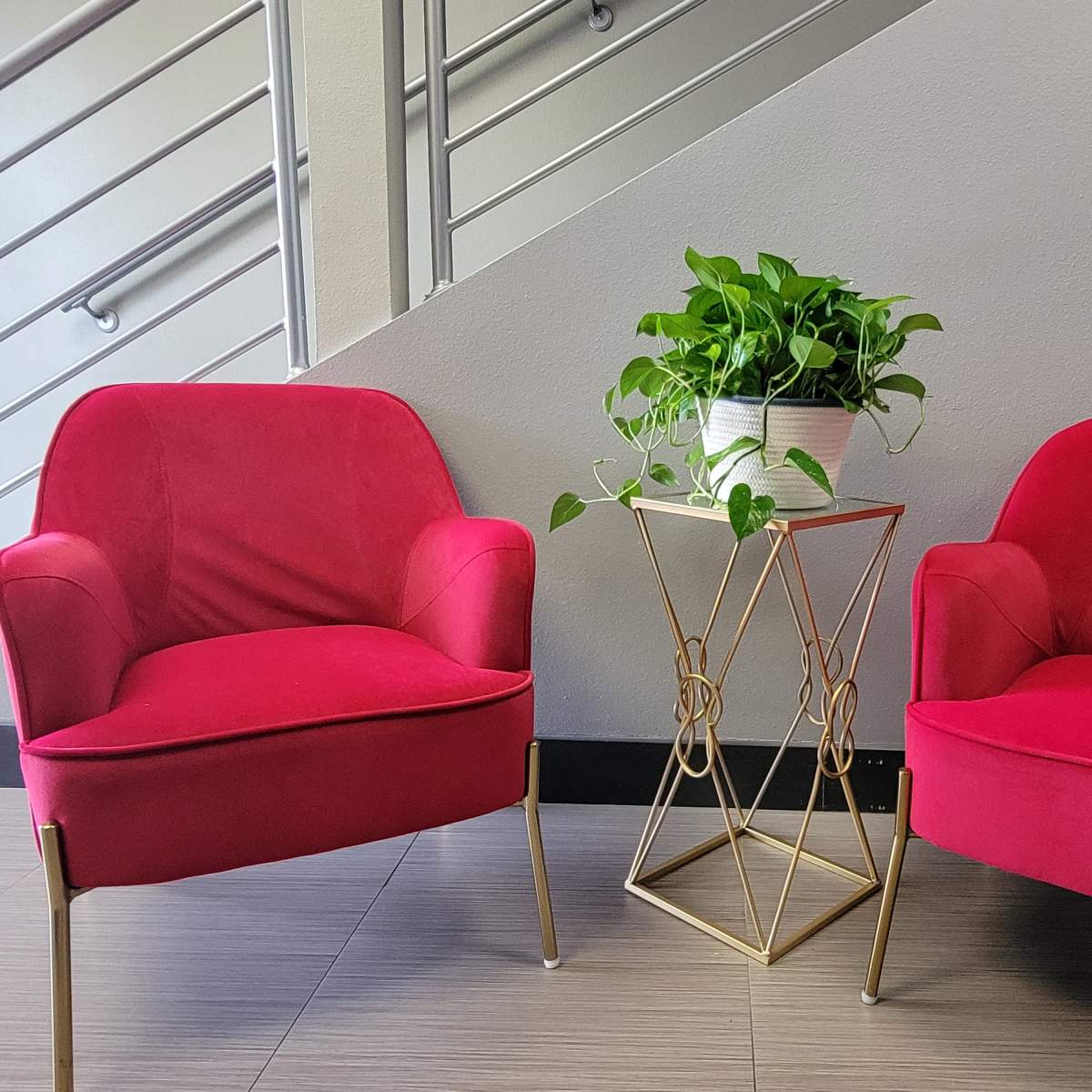 Two bright pink chairs are positioned on either side of a small gold metal table, which holds a potted plant, against a gray wall near a staircase.