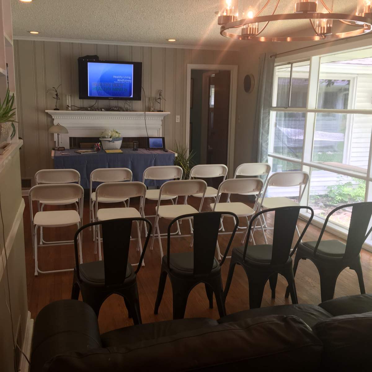 A spacious room is set up with rows of chairs facing a television displaying a presentation, with large windows allowing natural light to enter.