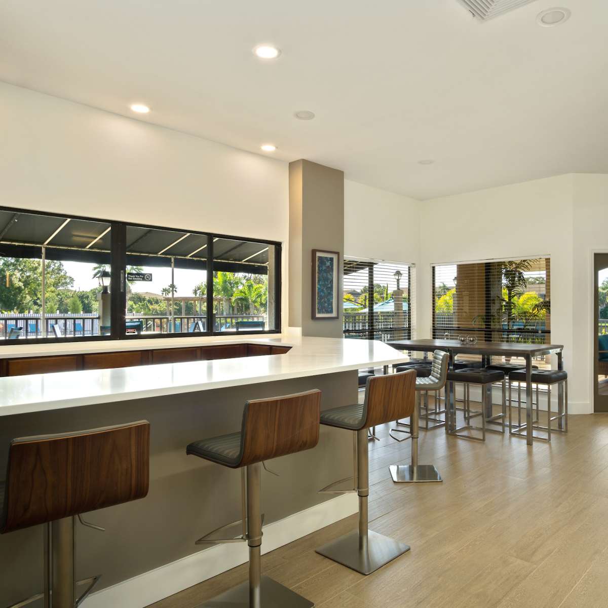 A modern interior space with a long countertop, bar stools, large windows overlooking greenery, and a seating area with additional tables and chairs.