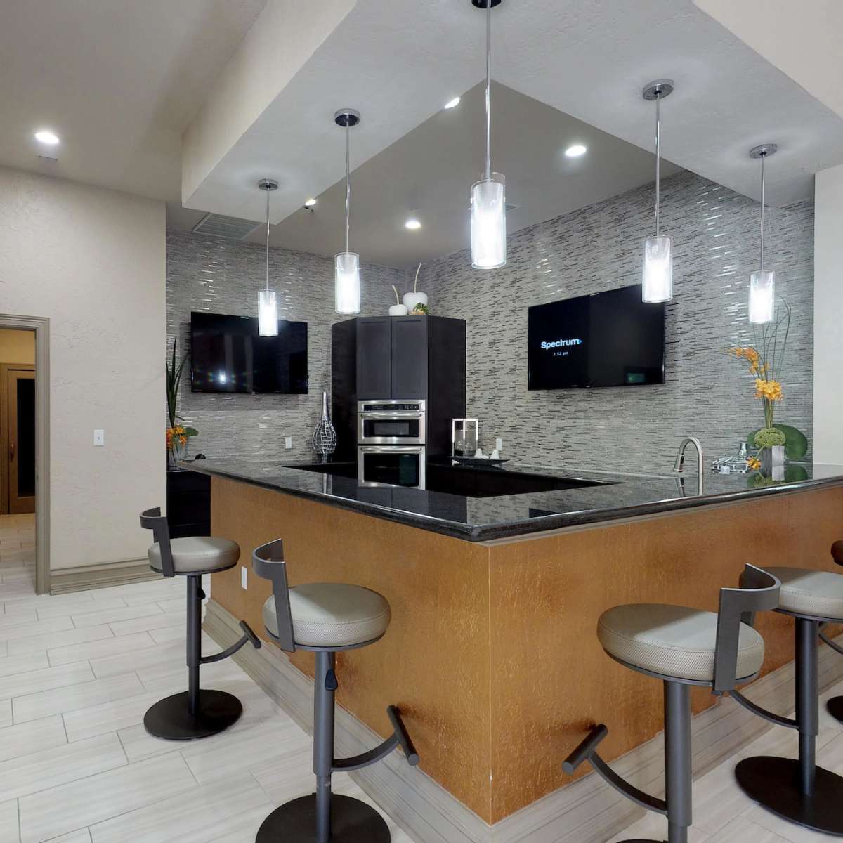 A modern kitchen with a bar counter, four stools, and two wall-mounted televisions.