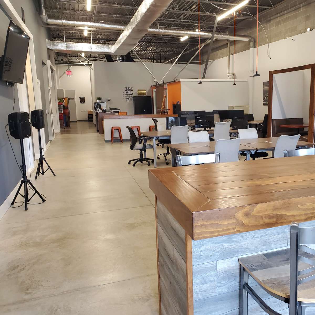 A modern co-working space with wooden tables, office chairs, and various workstations arranged across a spacious, well-lit interior.
