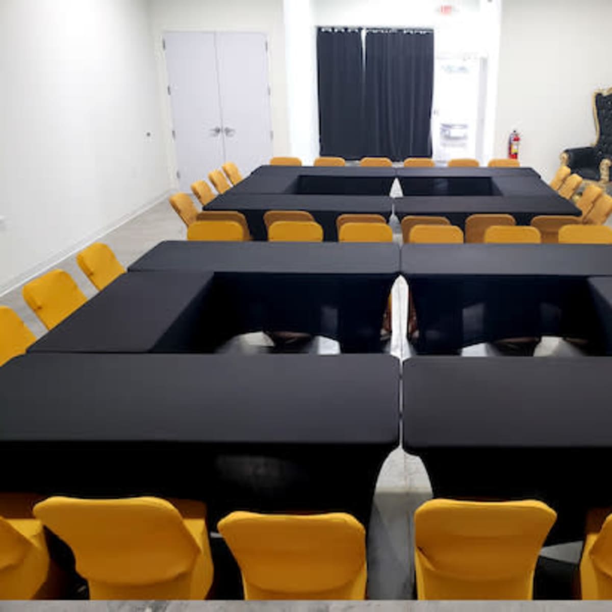 A rectangular arrangement of black tables and yellow chairs is set up in a meeting room with white walls and a decorative wall feature.