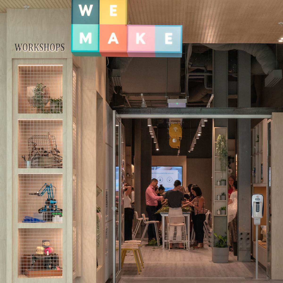 An entrance to a workshop space labeled "WE MAKE," featuring people gathered inside and various workstations along the walls.