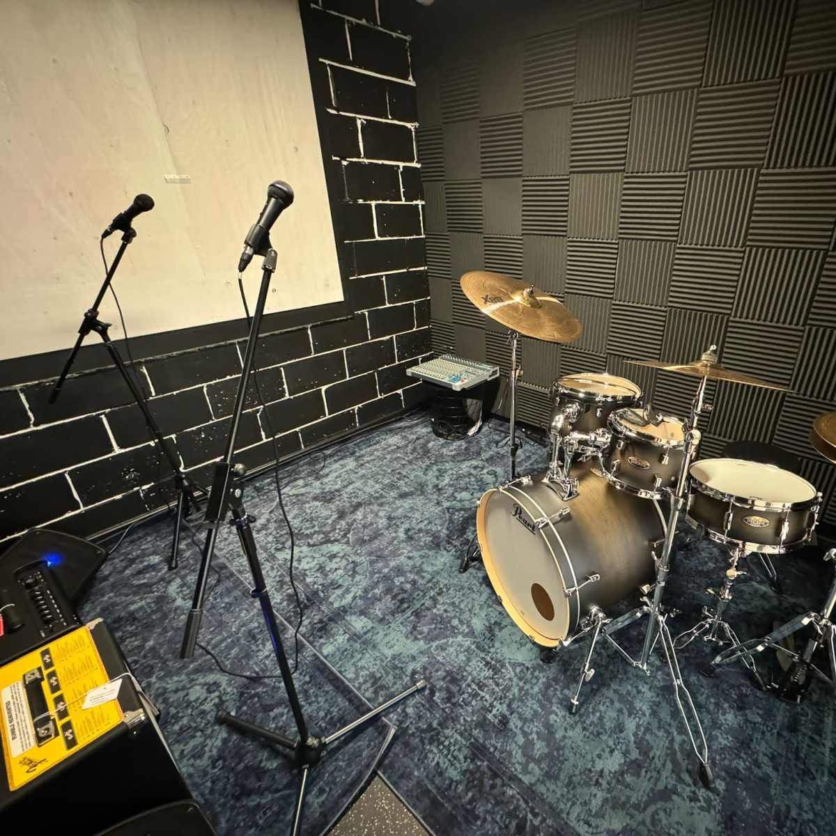 A rehearsal room featuring a drum set with cymbals, two microphones on stands, and a soundboard.