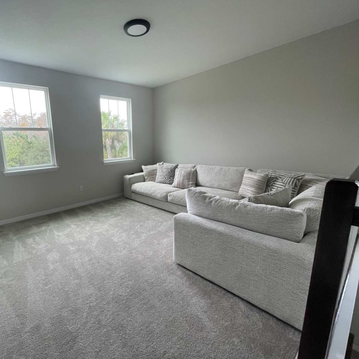 A light-filled living room features a large, plush sectional sofa and two windows overlooking greenery.