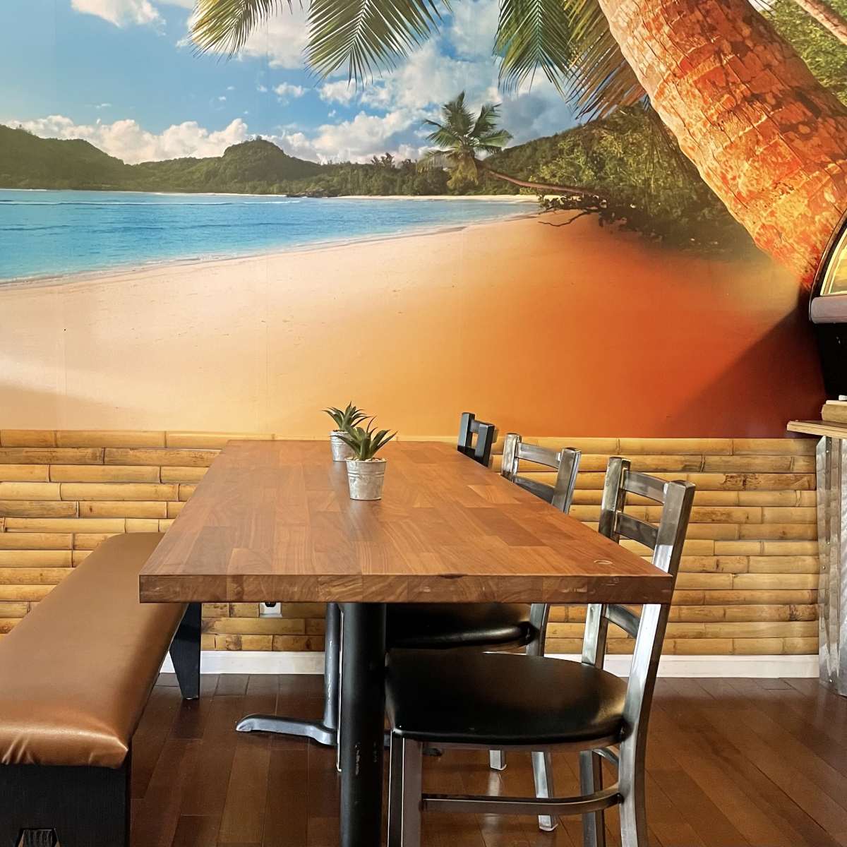 A wooden table and bench are set against a tropical beach mural with palm trees and a sandy shore.
