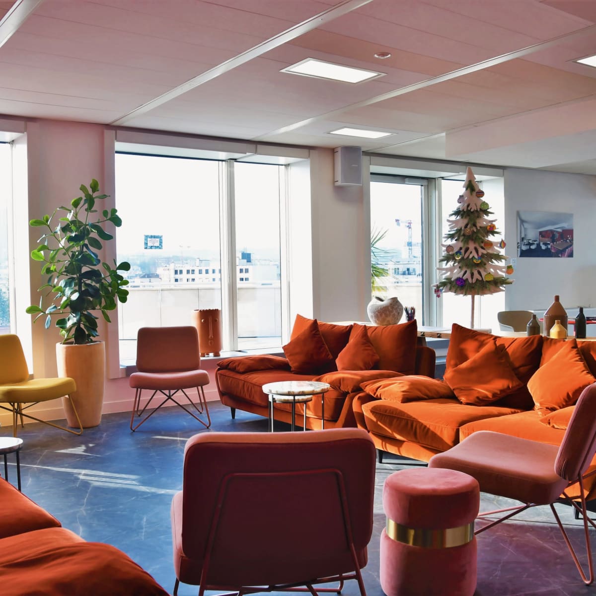 A modern lounge area with orange and pink sofas, small round tables, and large windows overlooking the city.