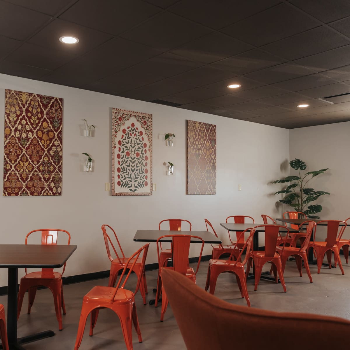 A cafe interior with orange metal chairs and tables, decorated walls featuring patterned artworks, and a few plants.