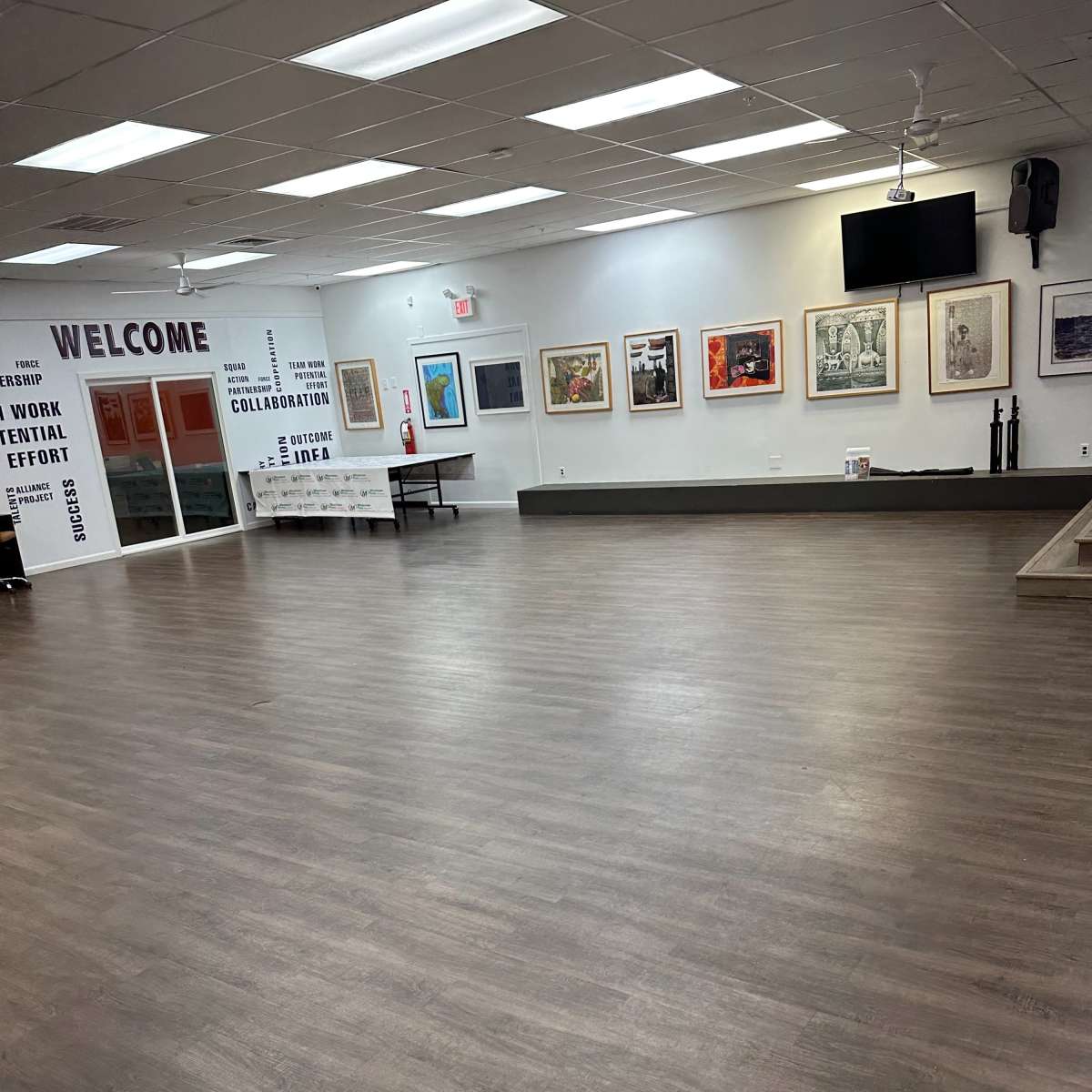 A spacious, empty room with wooden flooring, white walls adorned with framed artwork, and a welcome sign on one side.