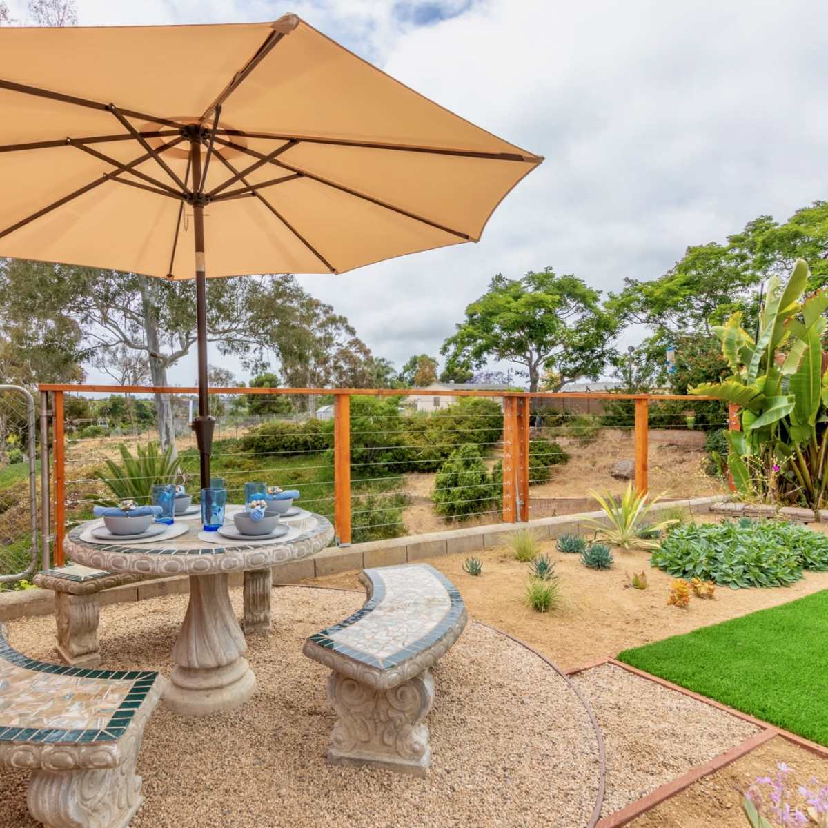 A stone table with matching curved benches is set under a large umbrella in a landscaped yard featuring greenery and decorative plants.