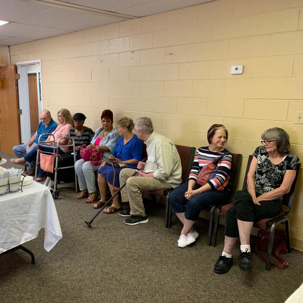 A group of people sits along a wall while a table with food and a cake is set up nearby in a community room.