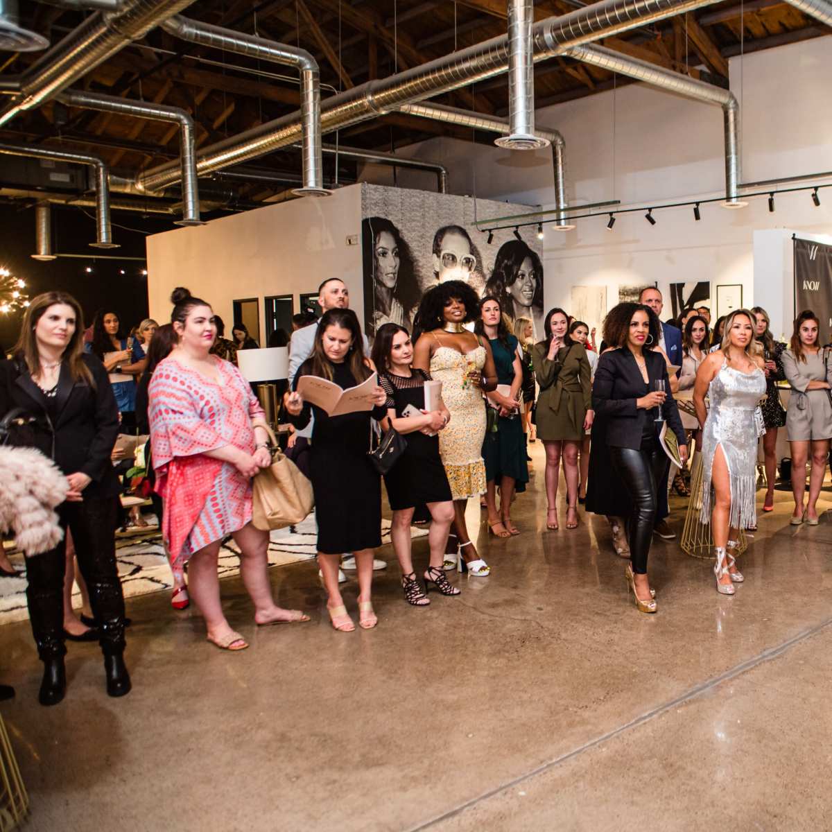 A crowd of diverse women gathers in a spacious gallery with artworks on the walls, while a speaker addresses the audience from the front.
