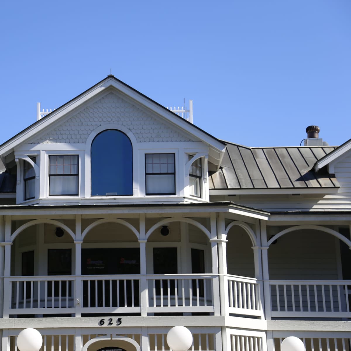 The upper portion of a multi-story house with a peaked roof, large windows, and a balcony surrounded by decorative white railings.