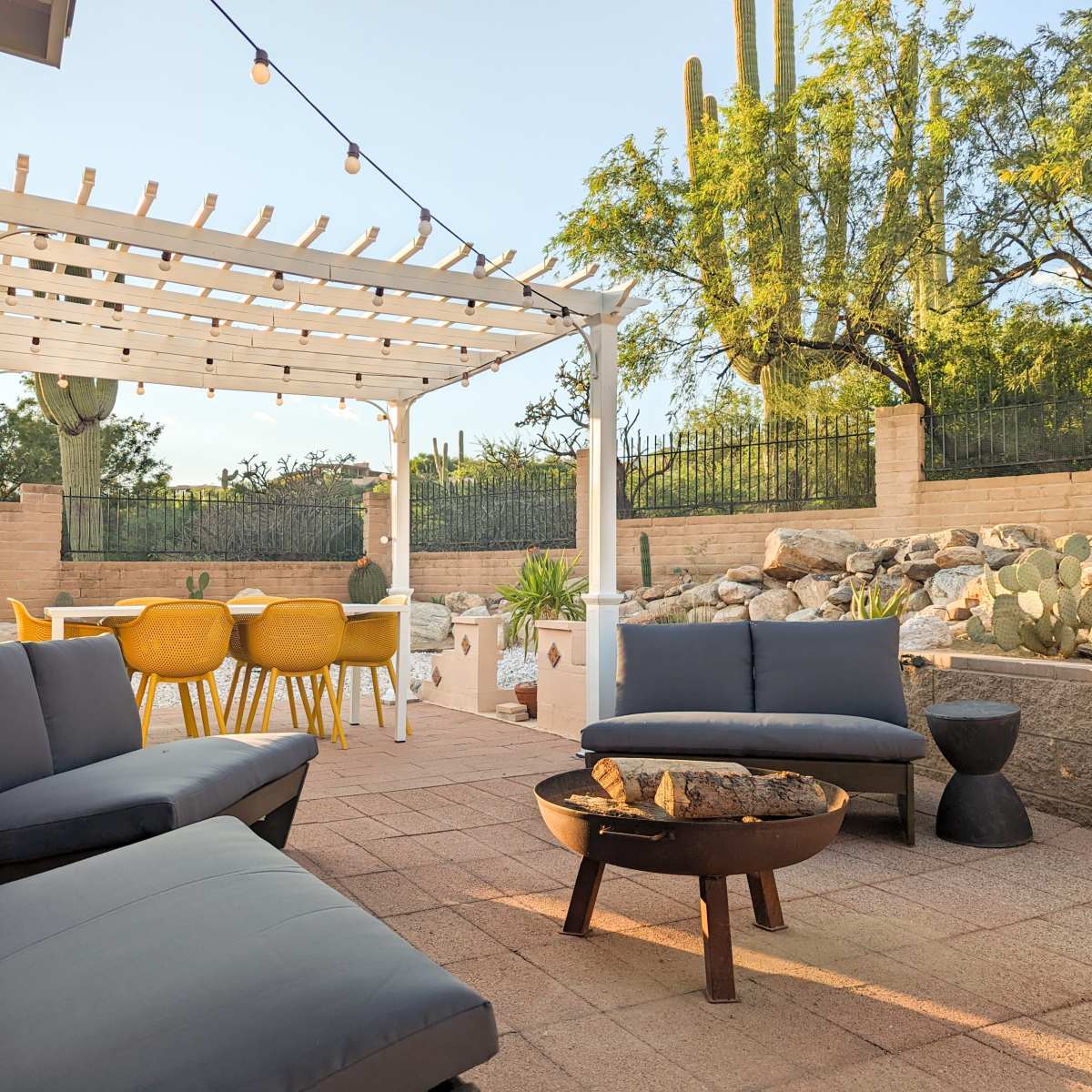 A patio area features modern seating, a fire pit, a dining table, and cacti in a landscaped yard under a clear sky.
