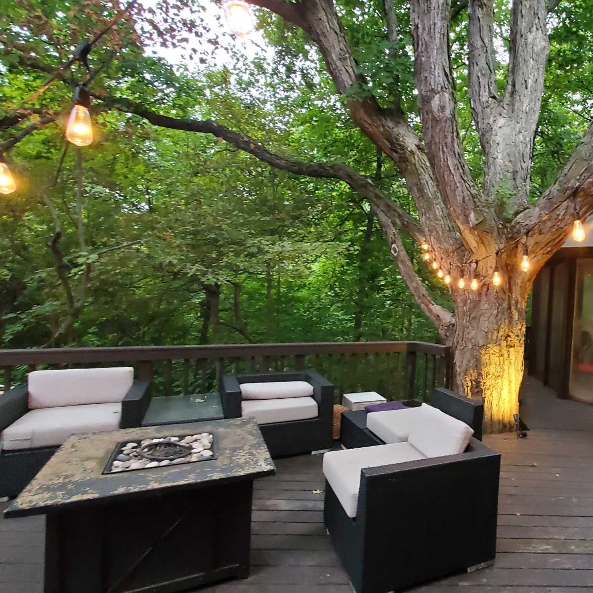 A wooden deck features comfortable seating, a fire pit, and a gazebo surrounded by trees illuminated with hanging lights.