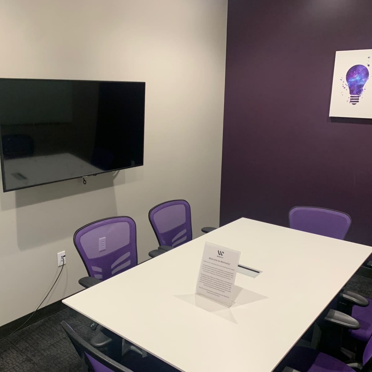 A modern meeting room features a white table surrounded by purple chairs and a wall-mounted television.