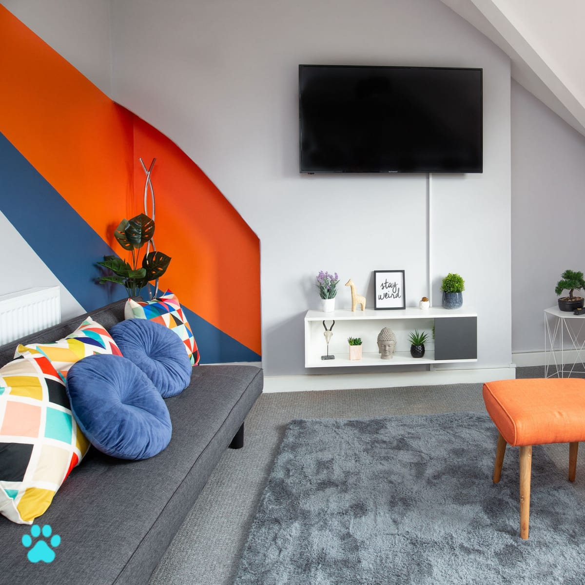The scene features a modern living room with a gray sofa, an orange accent chair, a white shelf mounted on the wall holding decorative items, and a large flat-screen TV.