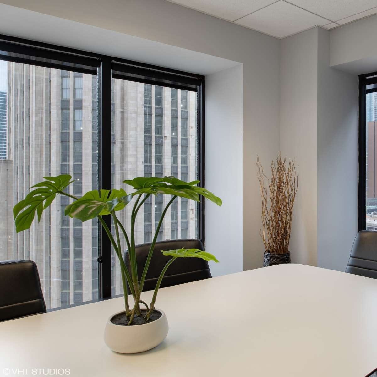 A conference room features a large white table, black chairs, and a potted plant near windows overlooking city skyscrapers and a river.