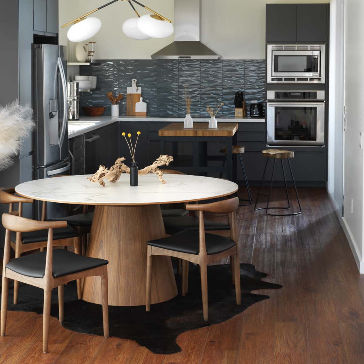 A modern kitchen and dining area features a round wooden table surrounded by four chairs, with a large vase of decorative branches on the table and a stylish cabinet in the background.