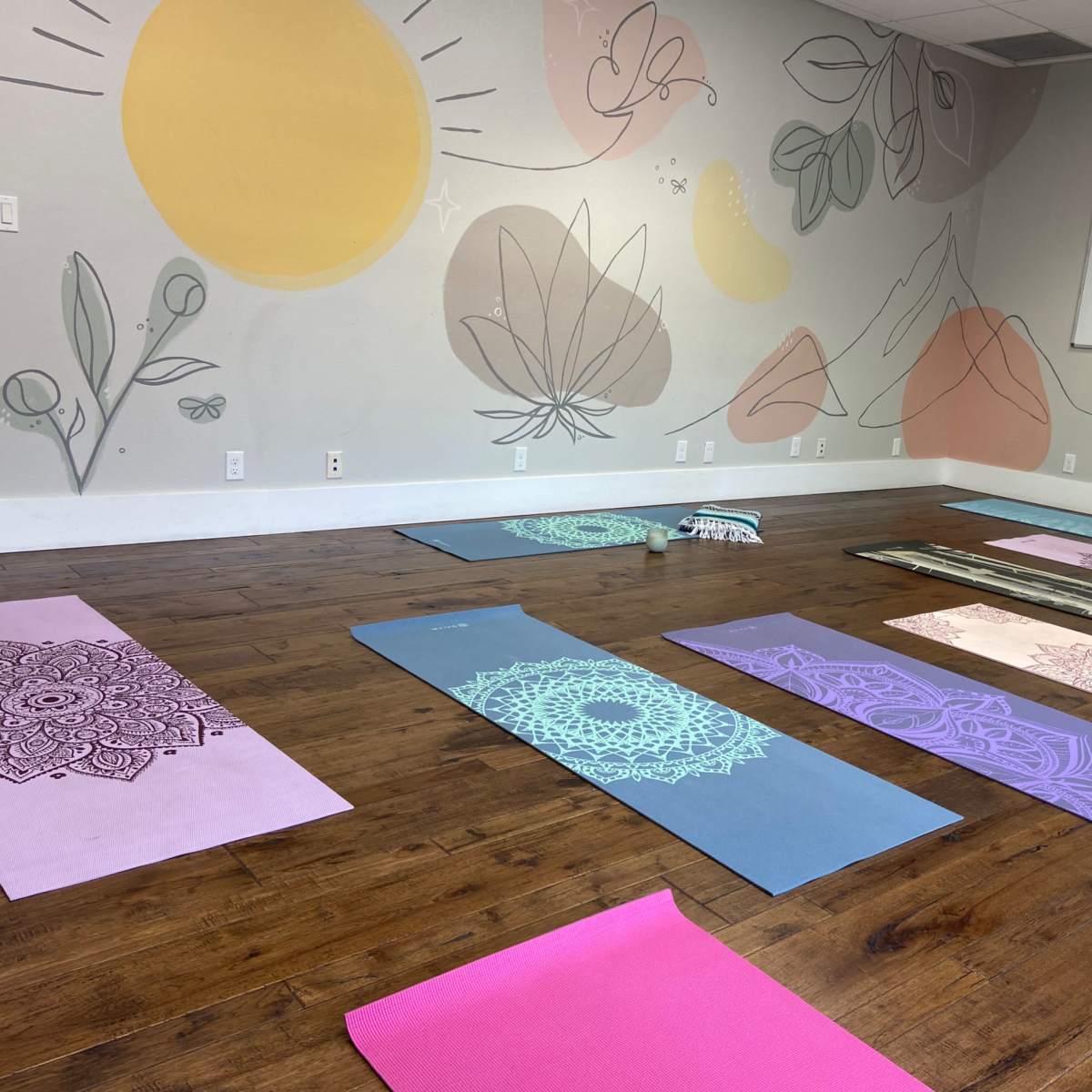 A yoga studio features multiple mats arranged on a wooden floor with decorative wall murals in soft colors.