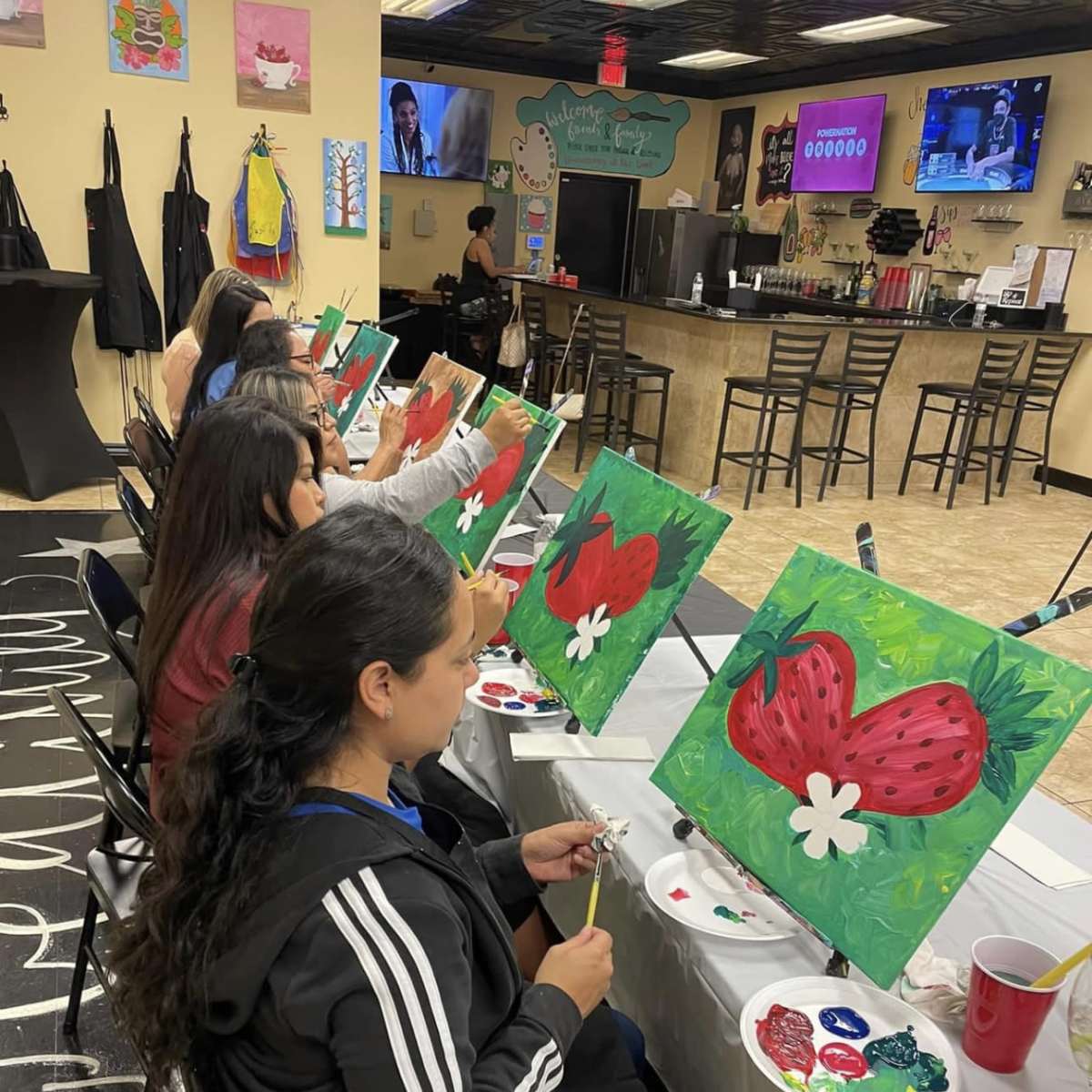 A group of individuals sits at tables in an art studio, painting strawberries on canvases.