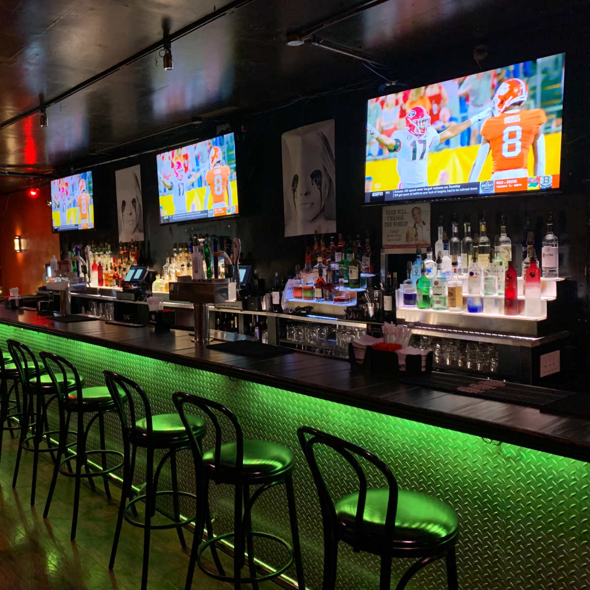 A dimly lit bar with a long green-lit counter, several bottles of liquor on display, two wall-mounted televisions showing a sports game, and a decorated Christmas tree in the corner.