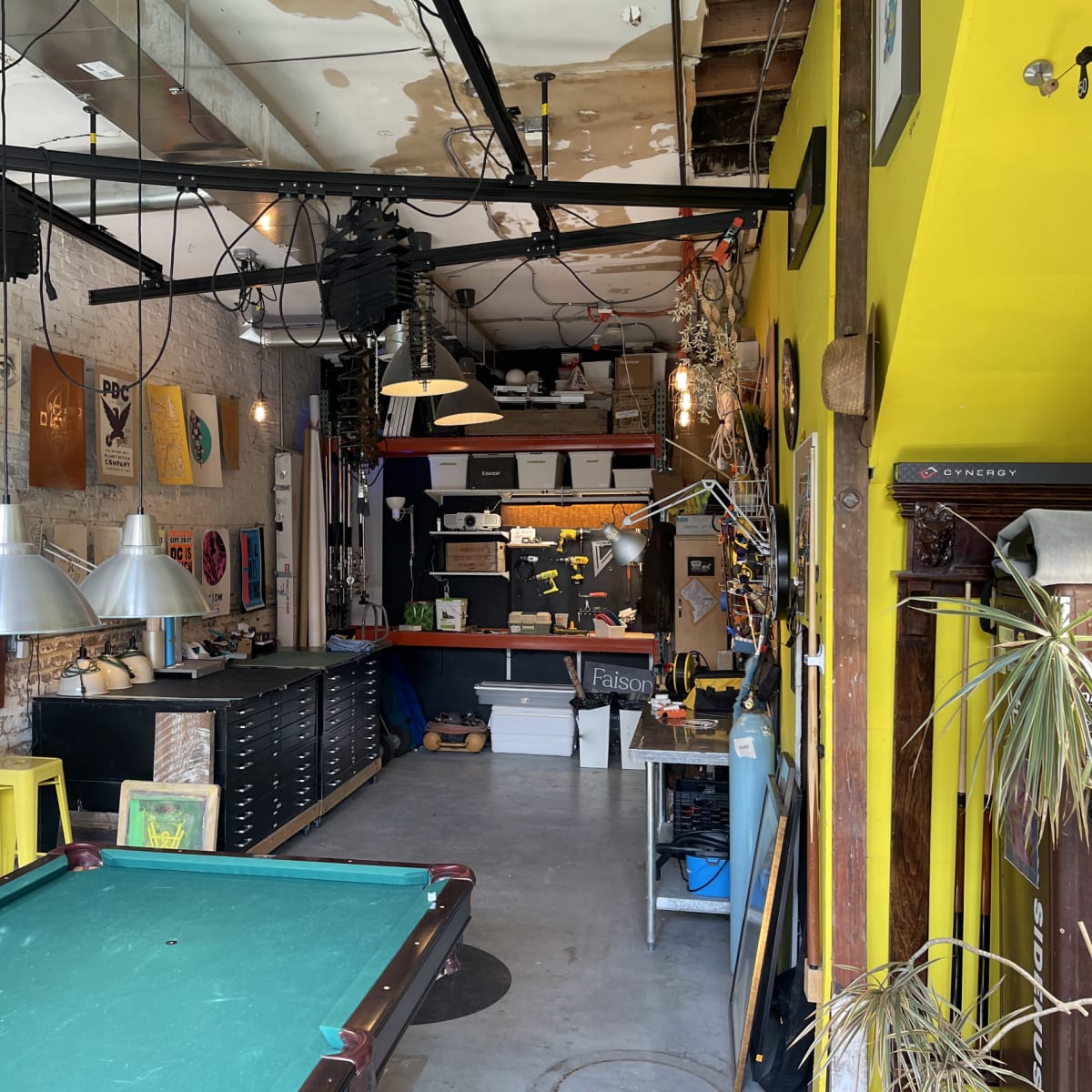 A well-lit workshop with a green pool table, yellow walls, and various tools and artwork displayed throughout the space.