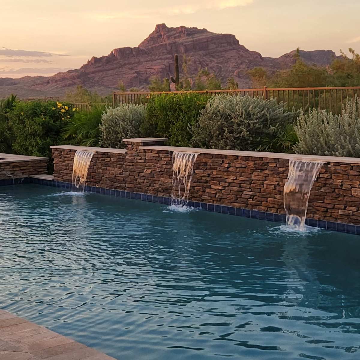 A swimming pool with stone waterfalls is situated in a landscaped area, framed by mountains in the background during sunset.