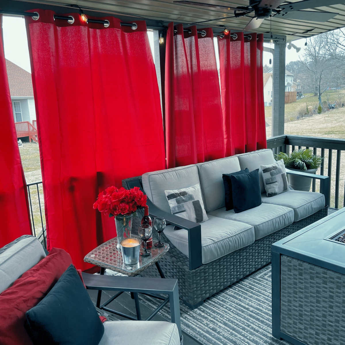 A covered outdoor seating area features a gray wicker sofa with red and black pillows, a glass-top fire pit table, red curtains, and a small table with a flower vase.
