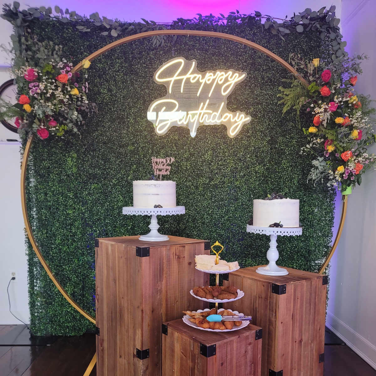 A birthday celebration setup with a neon "Happy Birthday" sign, two tiered cakes on pedestals, and a dessert display on wooden crates against a green backdrop.