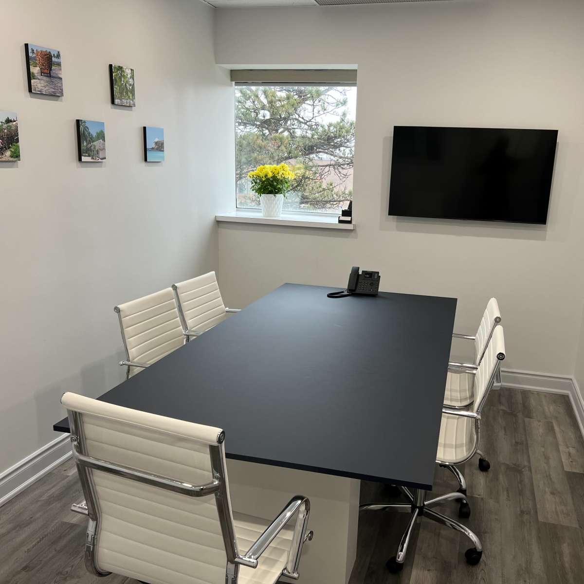 A conference room features a black table, four white chairs, a phone, and a television mounted on the wall, with framed photos arranged around the room and a window with flowers.