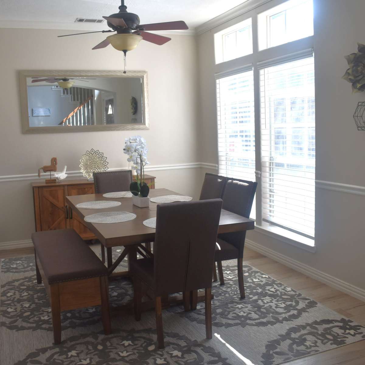 A dining area features a wooden table with upholstered chairs, a decorative mirror, and sunlight streaming through large windows.