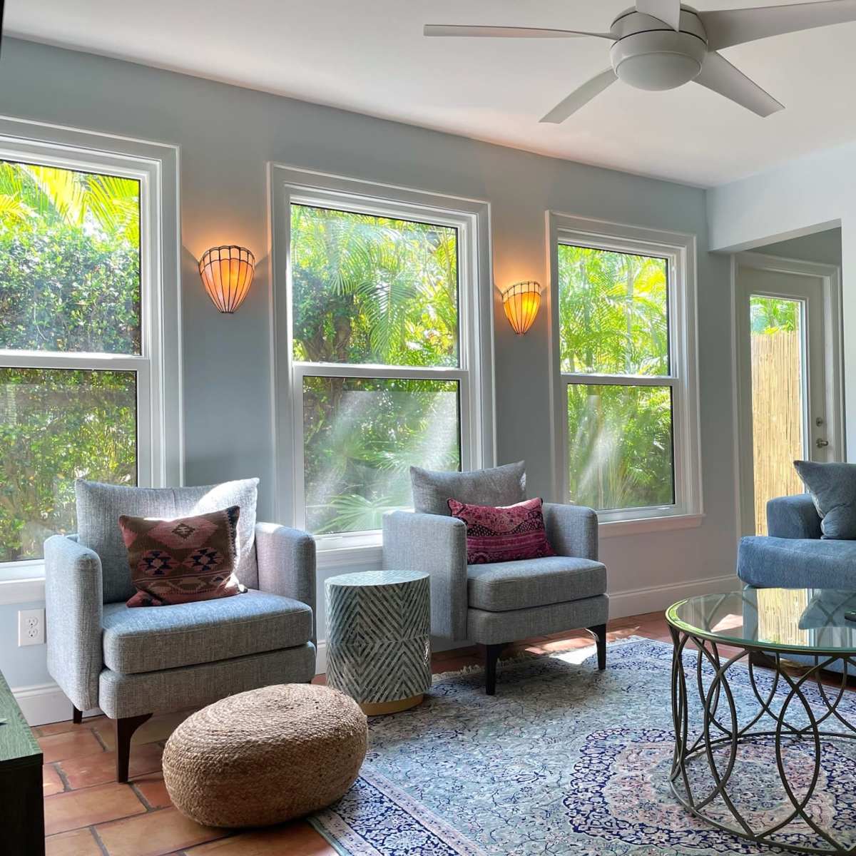 The living room features two gray armchairs, a glass coffee table on a patterned rug, and large windows allowing natural light to illuminate the space.