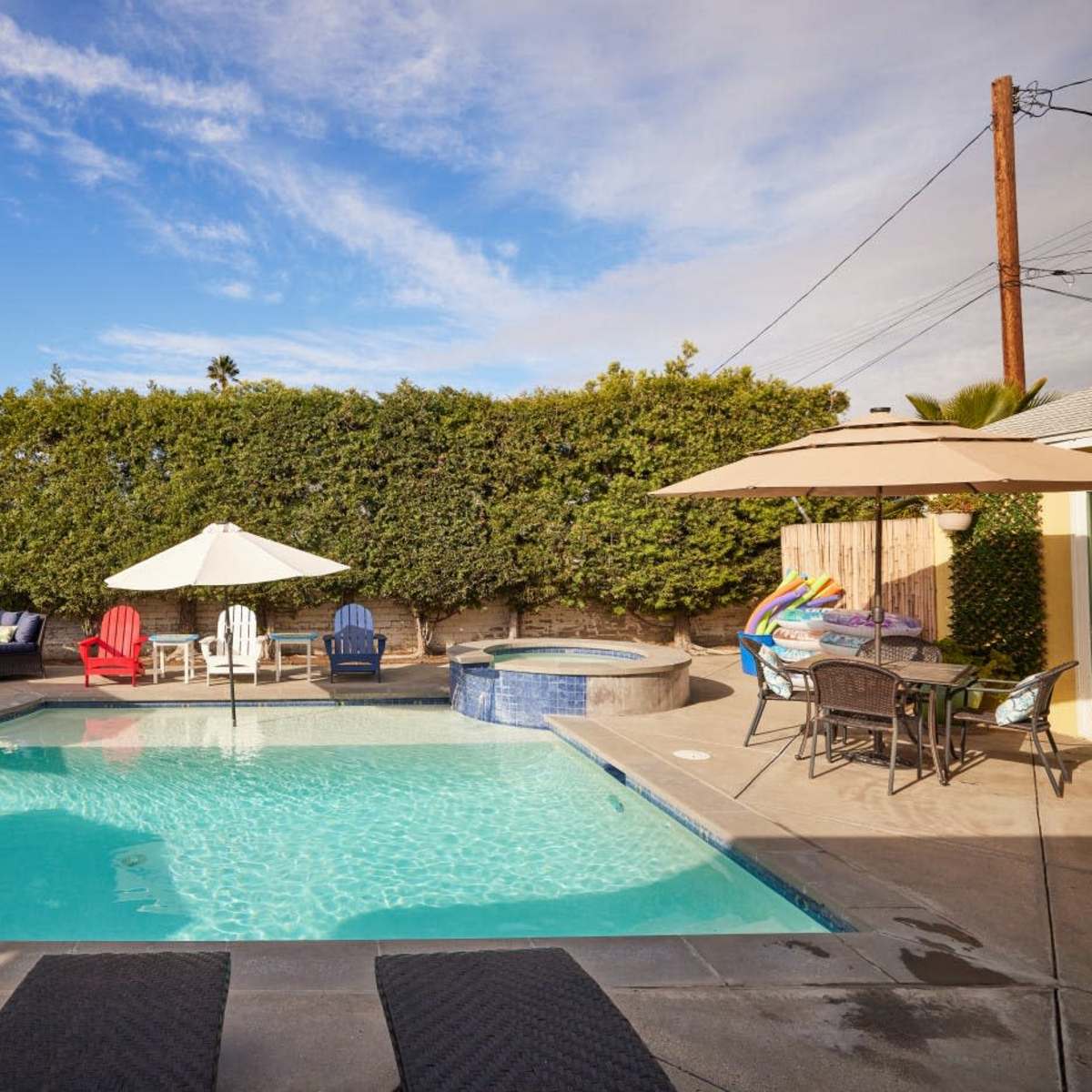A backyard pool area featuring a swimming pool, a hot tub, lounge chairs, and several umbrellas surrounded by landscaping.