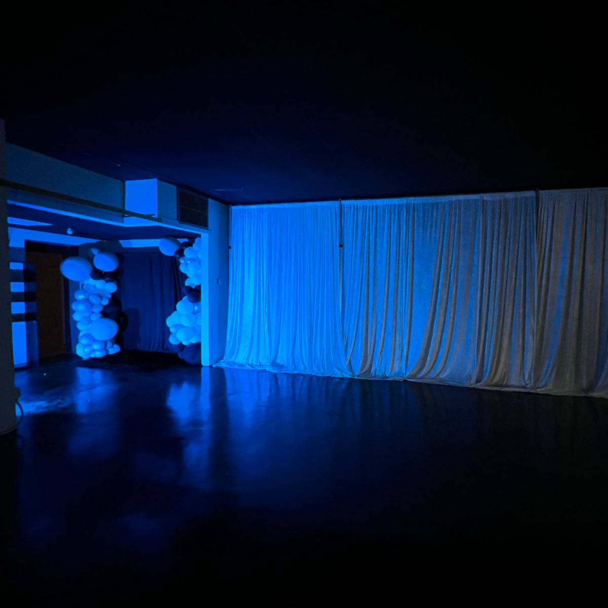 A dimly lit room with blue lighting, featuring a white curtain covering a wall and balloon decorations in the corner.