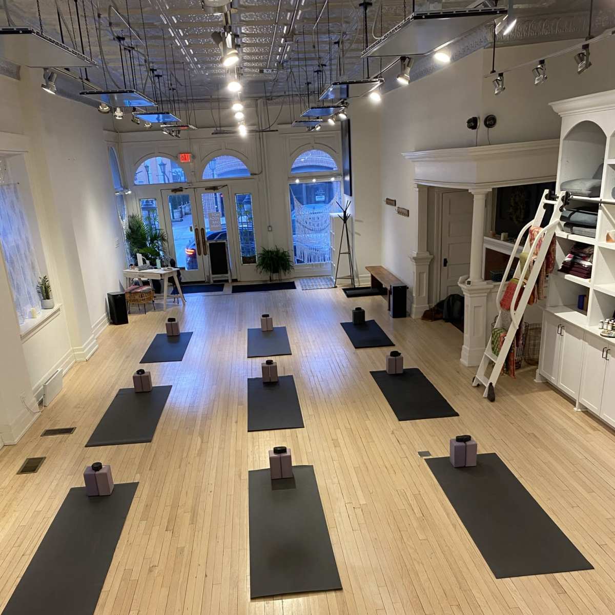 A spacious yoga studio with several black mats arranged in rows, each featuring a bolstered prop at the center, and large windows allowing natural light to enter.