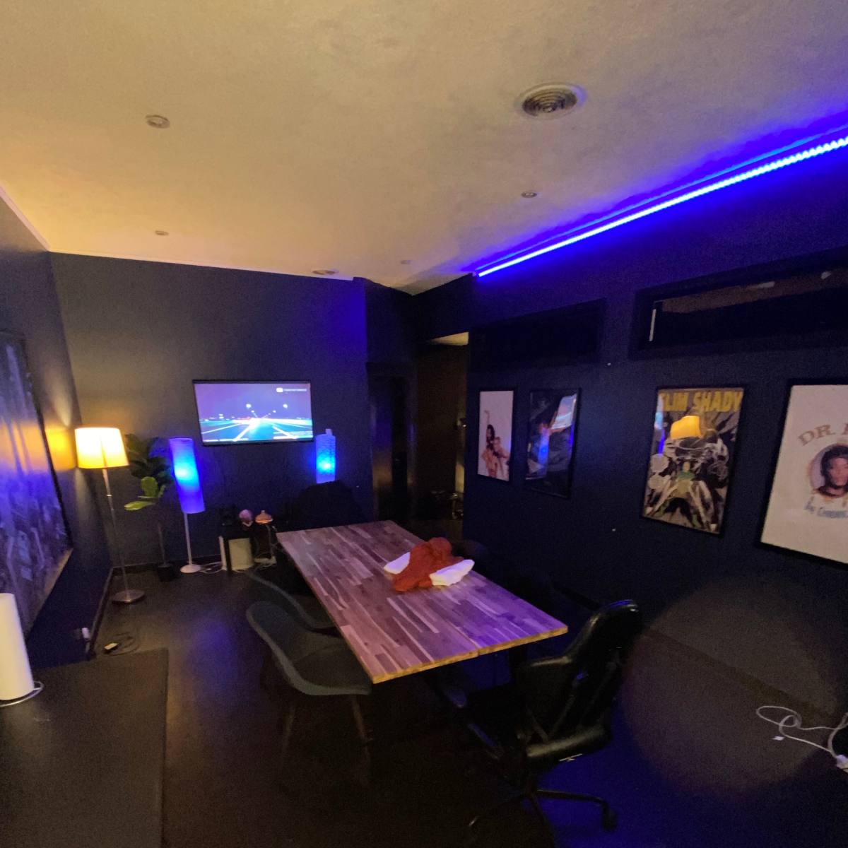 A dimly lit room with a large table, chairs, a wall-mounted TV, and framed posters on the walls.