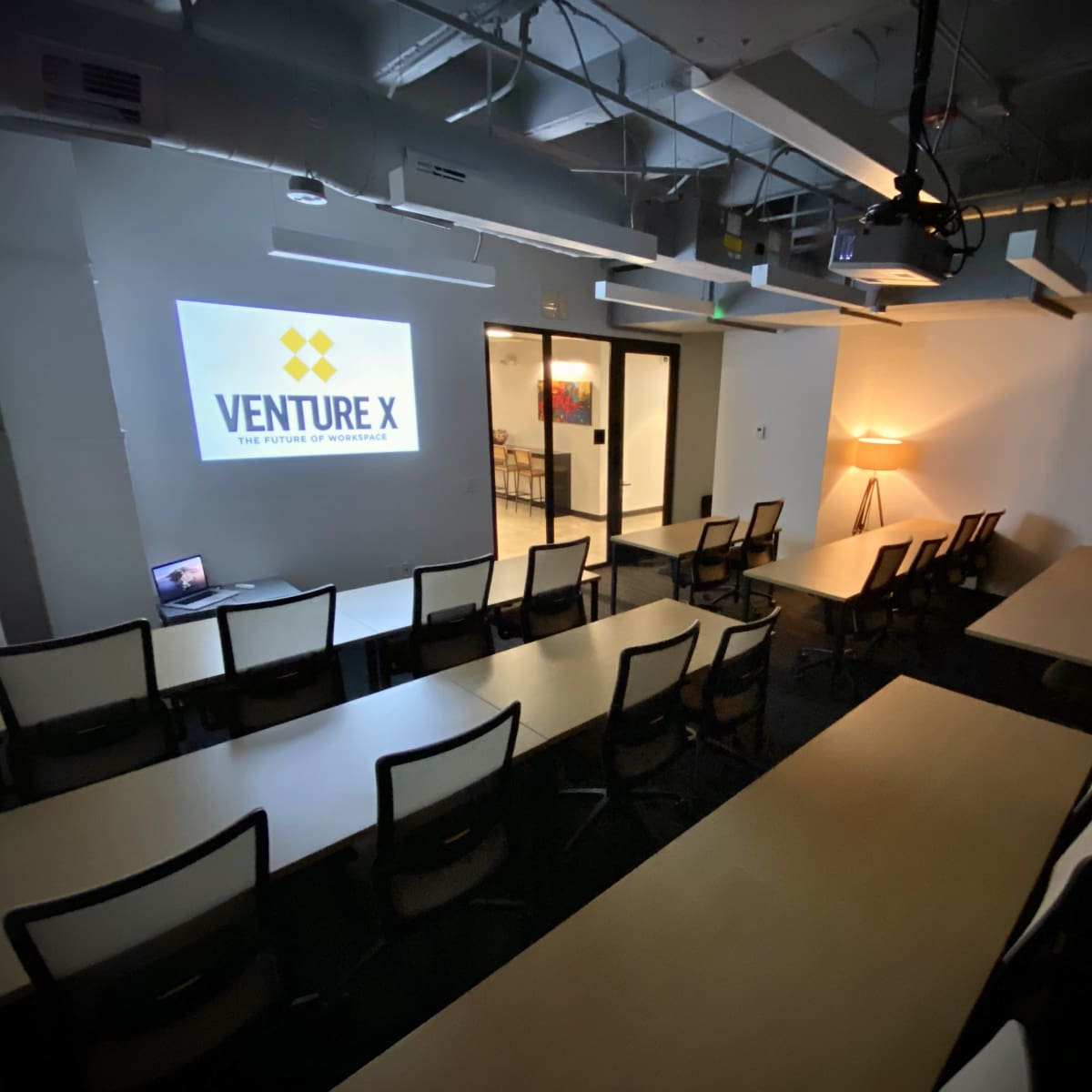 A conference room with rows of tables and chairs, featuring a projector displaying the "Venture X" logo on the wall.