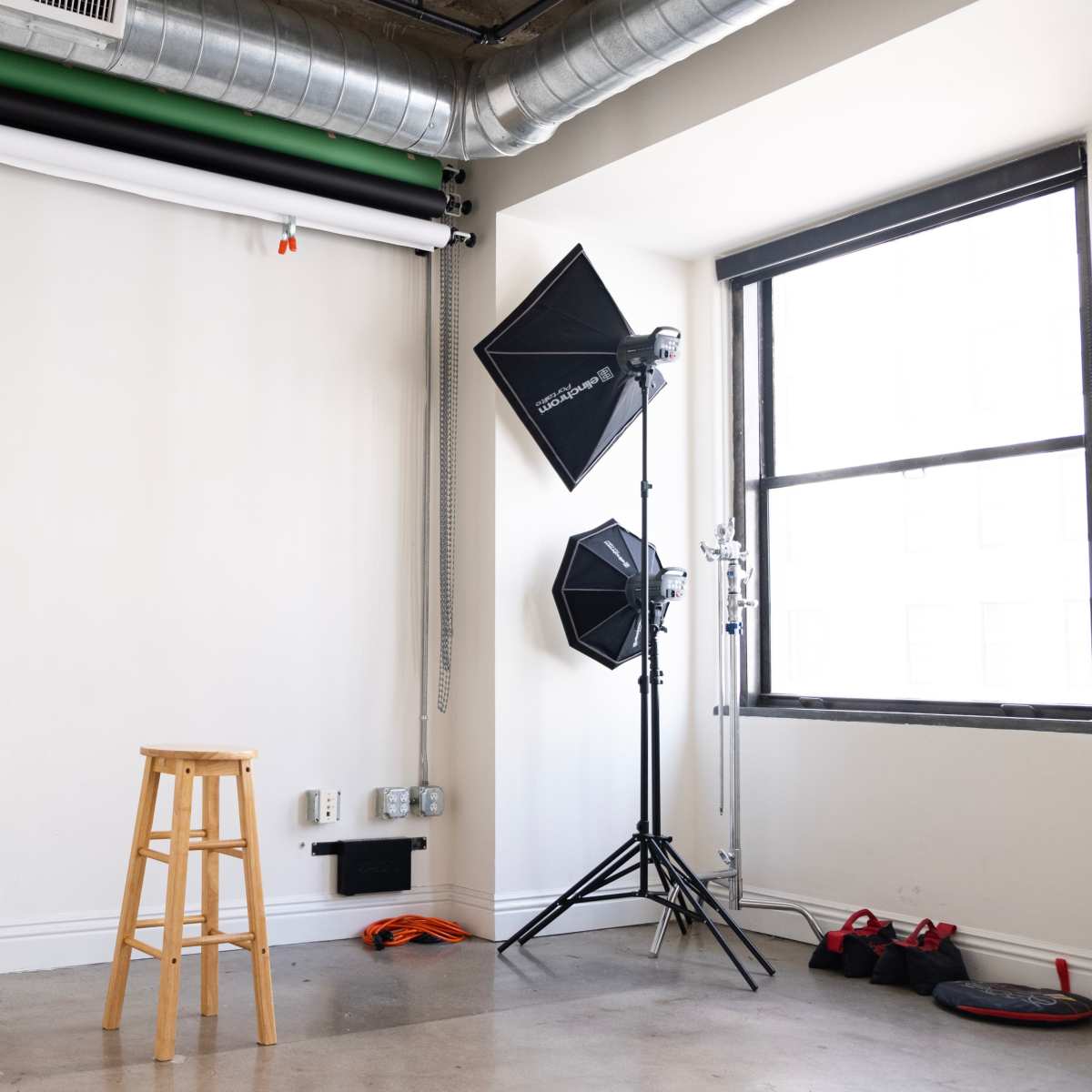 A photography studio with a wooden stool, lighting equipment, and a plain wall with a roll of backdrop.