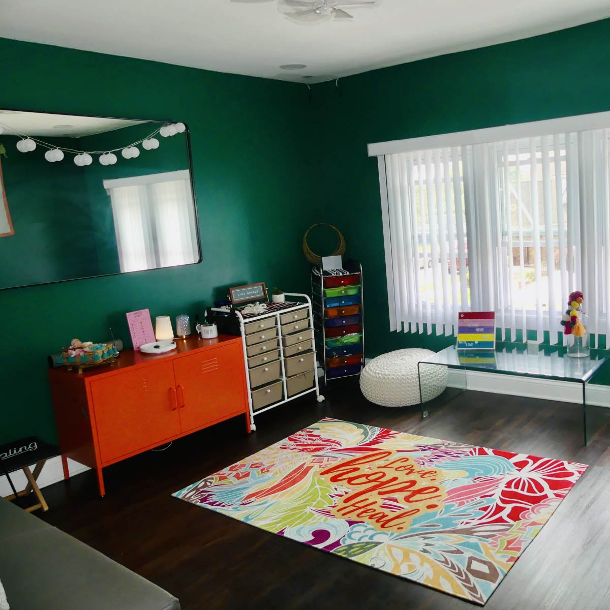 The room features green walls, a large window with white blinds, a colorful rug, and various furniture pieces including a glass coffee table and an orange cabinet.