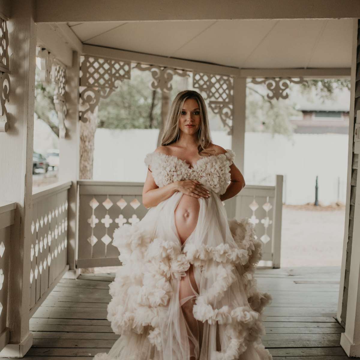 A pregnant woman stands indoors, wearing a flowing, ornate white dress with ruffles, and holds her hands on her belly.