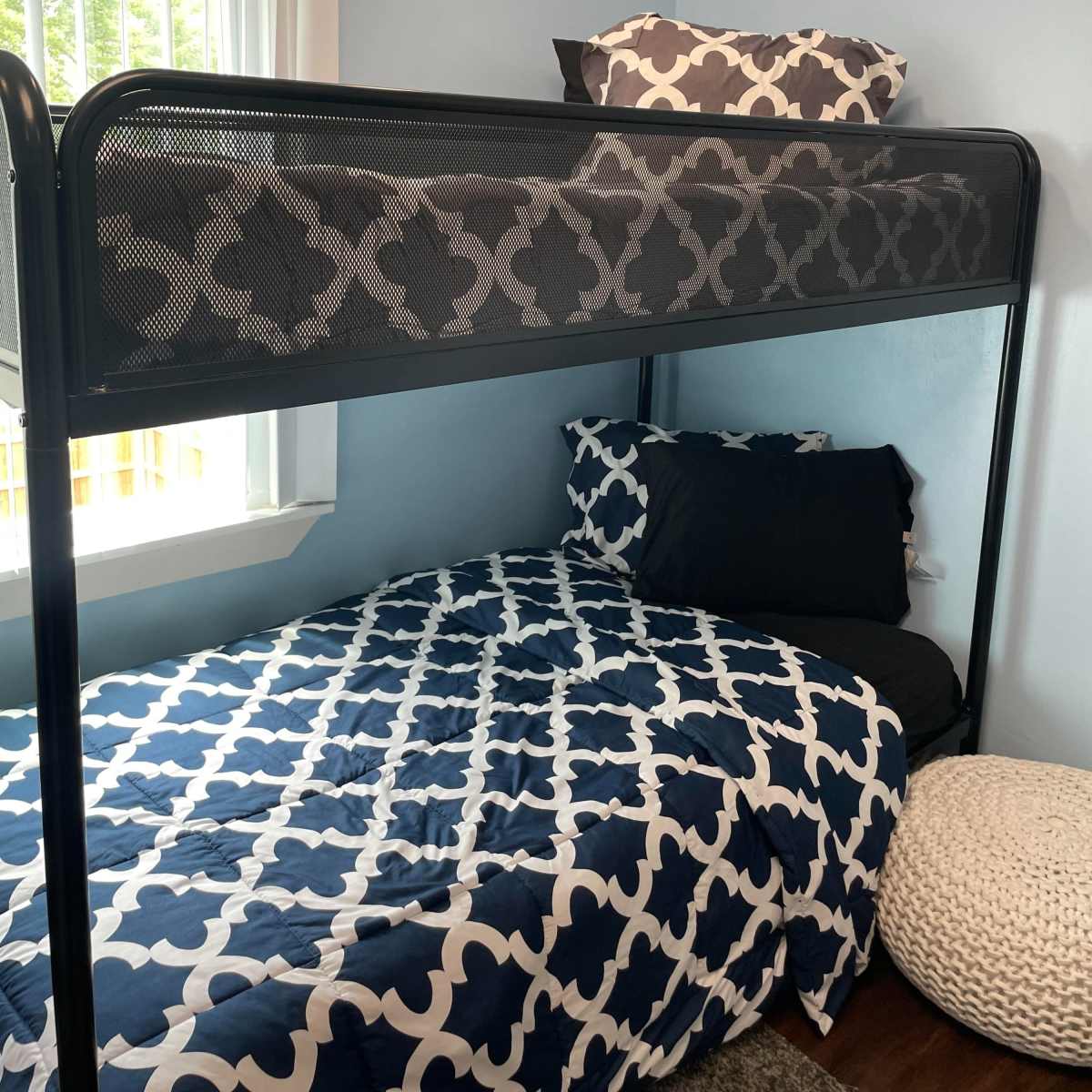 A bedroom with a lofted bed above a lower bed, both covered with a patterned blue and white bedspread, and a round pouf on the floor.