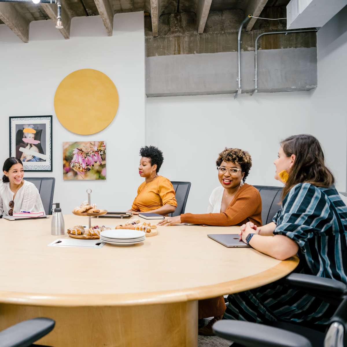 A group of six people is seated around a large circular table in a bright meeting room while a woman presents at the front of the room.