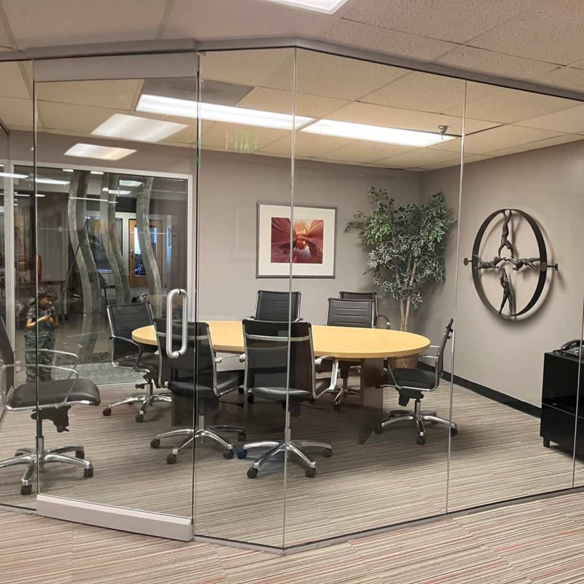 A glass-walled conference room with a circular table surrounded by black chairs, a plant in one corner, and a wall-mounted decoration.